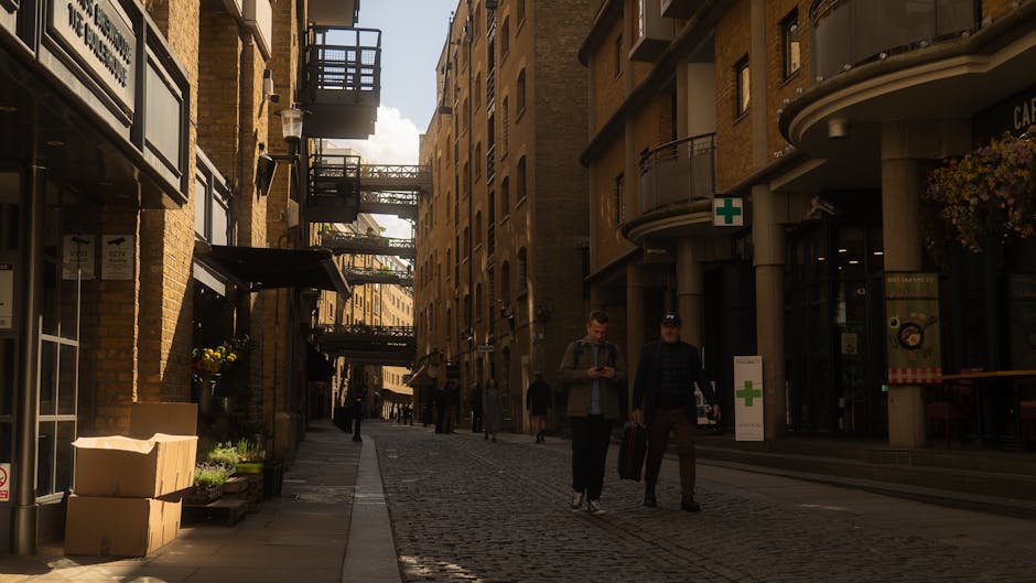 A narrow cobblestone street in an urban area, lined with multi-storey brick and concrete buildings on both sides, with fire escape balconies and external walkways connecting the upper floors. On the left side, there are cardboard boxes, some stacked and others placed on the ground near a storefront with potted plants and outdoor signage. Two men are walking along the street; one is holding a smartphone, and the other is carrying a wheeled suitcase, indicating they may be involved in a home relocation or moving process. The scene is lit by natural daylight, with a partly cloudy sky visible overhead. In the background, other pedestrians are seen walking further down the street. The environment and the presence of moving equipment directly relate to logistics and packing activities associated with house removals, as managed by companies such as Man and Van Finchley, fitting within the context of a Finchley Central removals guide for narrow streets.