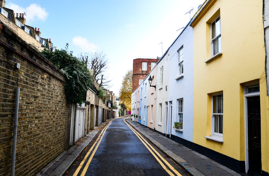A narrow residential street during daylight showing a row of terraced houses with painted facades in pastel yellow, pale blue, and white, positioned directly adjacent to the pavement. The pavement is made of concrete slabs, and the street surface is asphalt with double yellow lines along the curb. On the left side, there is an aged brick wall with some plant growth, and small garage doors are visible along this wall. The buildings have modern double-glazed windows, some with flower boxes underneath. The scene appears quiet, with no vehicles or people present, and the area is illuminated by natural sunlight with partly cloudy skies overhead. This setting exemplifies typical local streets where house removals and furniture transport services by companies like Man and Van Finchley often take place during home relocations or packing and moving processes across Finchley.