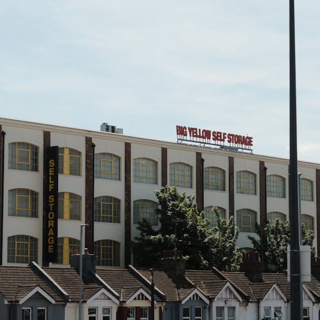 Exterior view of a multi-storey industrial building with a white facade and large, arched yellow-framed windows, labeled 'Self Storage' with a vertical sign on the left side. A red sign on the roof reads 'Big Yellow Self Storage.' In the foreground, there are small residential houses with pitched roofs and chimneys, and a few leafy trees partially obscuring the lower part of the building. The scene is captured during daylight with a partly cloudy sky, and a streetlamp pole is visible on the right side. This image illustrates the typical environment where professional house removals and furniture transport services, such as those offered by Man and Van Finchley, could facilitate home relocation or packing and moving processes involving storage facilities.