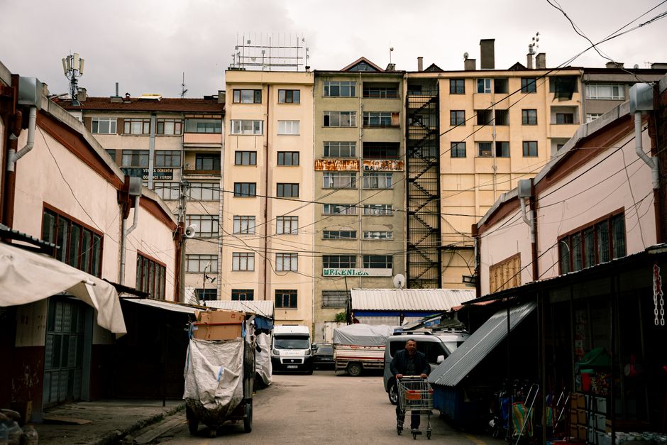 The image depicts a narrow urban street with a large, multi-storey residential building in the background, featuring multiple windows, external staircases, and balconies. In the foreground, there are several small shops or market stalls with closed shutters, some covered with fabric awnings. A man is visible pushing a shopping trolley along the street, which is lined with parked vans and trucks used for home relocation and furniture transport. Materials such as cardboard boxes, wrapped furniture, and packing supplies are evident near the vehicles, indicating ongoing packing and moving activities associated with house removals. Overhead, multiple electrical wires cross the scene, and the overall environment appears to be a typical city area used for local packing, loading, and transport operations connected to home moving services offered by Man and Van Finchley, as seen on the page about North Finchley man and van tips for flats above shops.