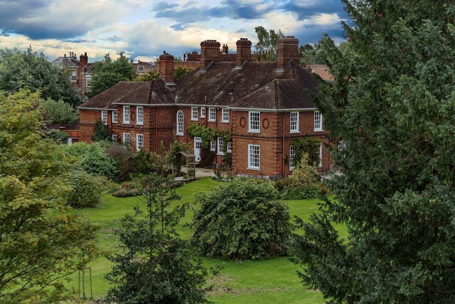 A large red-brick building with multiple tall chimneys and white-framed windows, surrounded by lush green trees and well-maintained gardens, situated within a residential area. The house appears to be an older, historic property, with a pathway and small garden features visible near the entrance. In the foreground, a dense collection of various trees and shrubs partially obscures the view of the building. The sky above is cloudy with patches of blue, indicating a partly cloudy day. This setting reflects the type of property that might be involved in a home relocation or furniture transport process, as part of professional removals services by Man and Van Finchley.