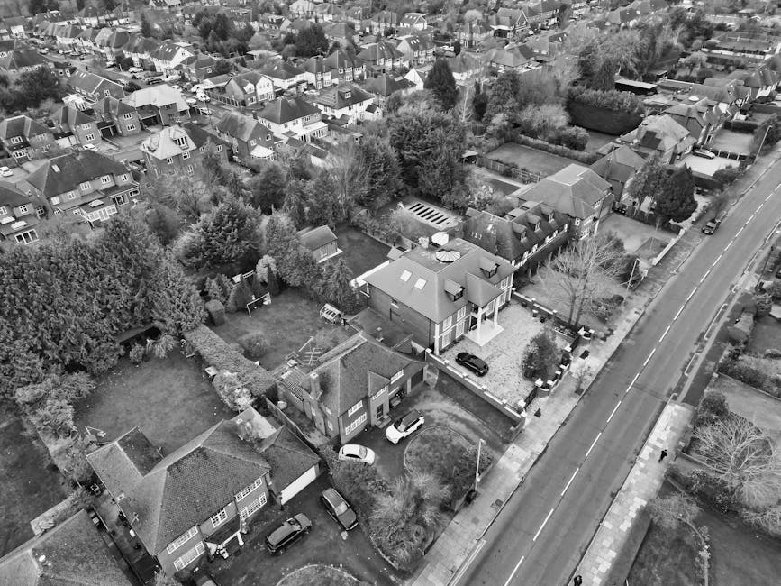 An aerial view of a residential area showing multiple houses with various roof styles and garden layouts, taken during daylight. In the foreground, a large detached house features a gravel driveway with several parked cars and a small front garden with trees and shrubs. Behind this property, there is a backyard enclosed by a fence, containing lawn space, trees, and a small outbuilding. Adjacent to the properties, a main road runs along the right side of the image, with parked vehicles and a sidewalk. The surrounding neighborhood displays a mixture of terraced and semi-detached houses, some with visible gardens and driveways, as well as tree-lined streets and patches of greenery. This detailed outdoor scene reflects a typical home environment that could be involved in a furniture transport or house removals process, as coordinated by professional services like Man and Van Finchley for home relocation and packing activities.