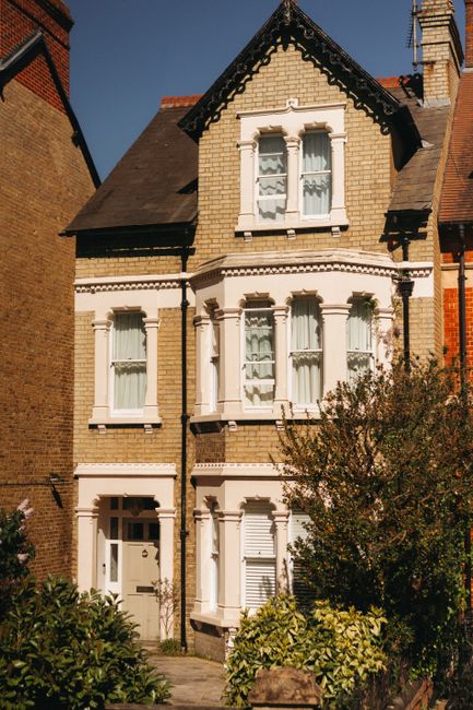A large red-brick building with multiple tall chimneys and white-framed windows, surrounded by lush green trees and well-maintained gardens, situated within a residential area. The house appears to be an older, historic property, with a pathway and small garden features visible near the entrance. In the foreground, a dense collection of various trees and shrubs partially obscures the view of the building. The sky above is cloudy with patches of blue, indicating a partly cloudy day. This setting reflects the type of property that might be involved in a home relocation or furniture transport process, as part of professional removals services by Man and Van Finchley.